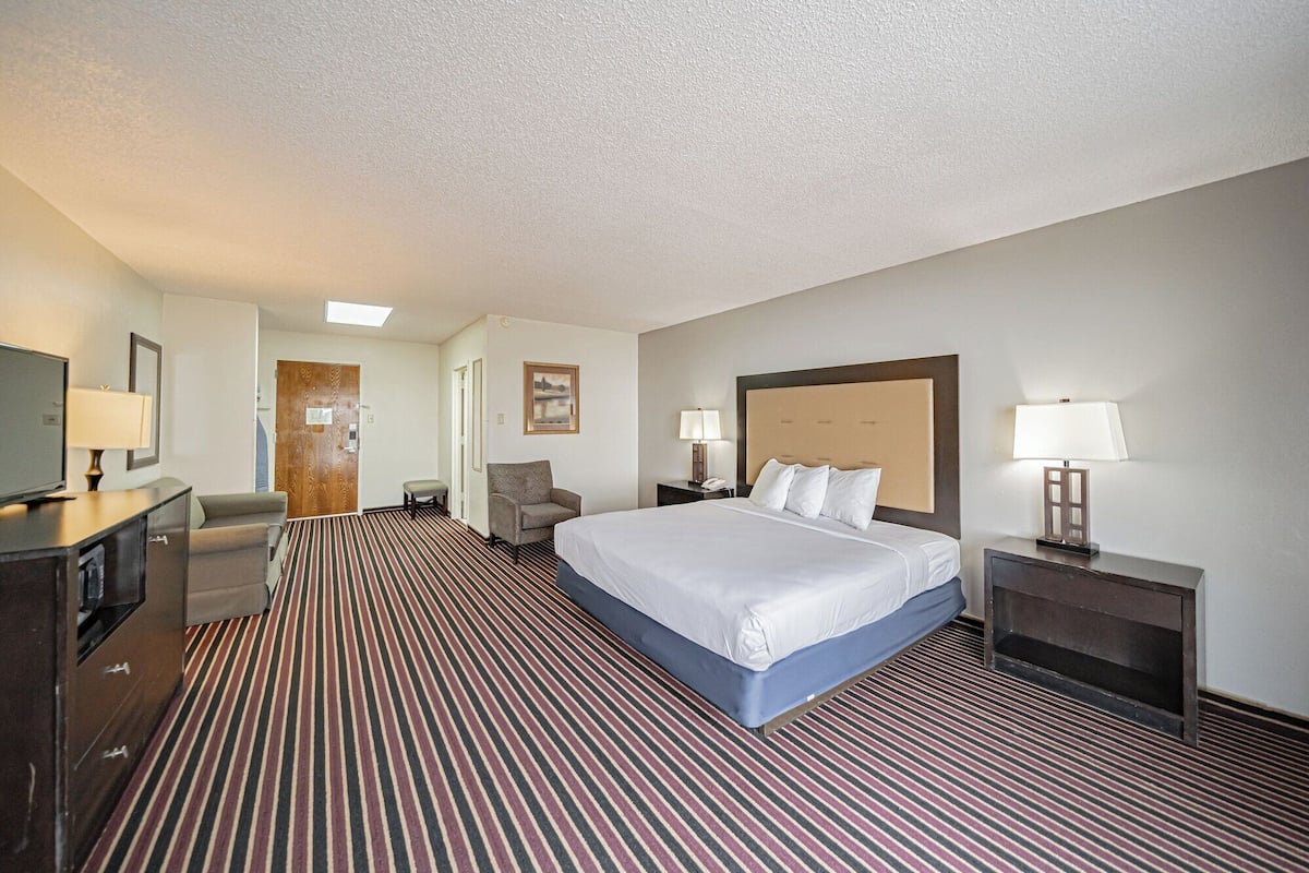 A hotel room with a large bed, a desk with a chair, a television, and a door leading to another room.