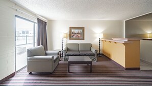 A hotel room with a sofa, armchair, and a small table. There is a painting on the wall and a window with curtains.