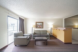 A hotel room with a sofa, armchair, and a small table. There is a painting on the wall and a window with curtains.