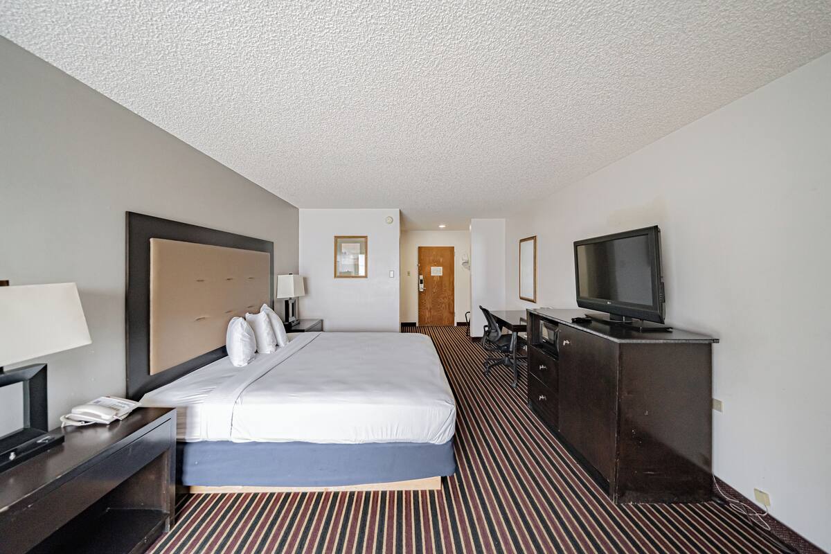 A hotel room with a large bed, a desk with a lamp, a television on a dresser, and a door leading to another room.