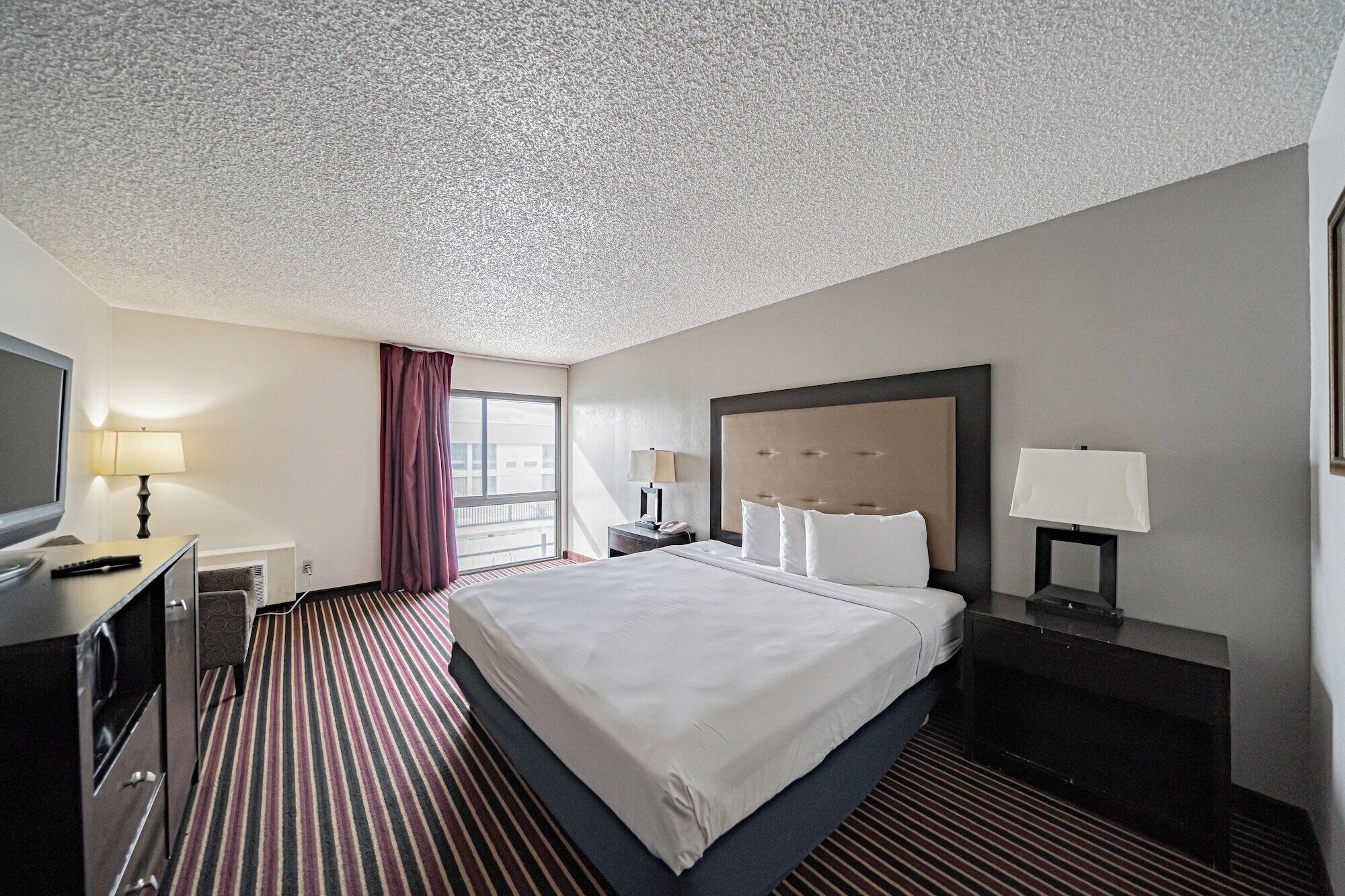 Habitación de hotel con una cama grande, un escritorio con lámpara, un televisor y una ventana con cortinas.