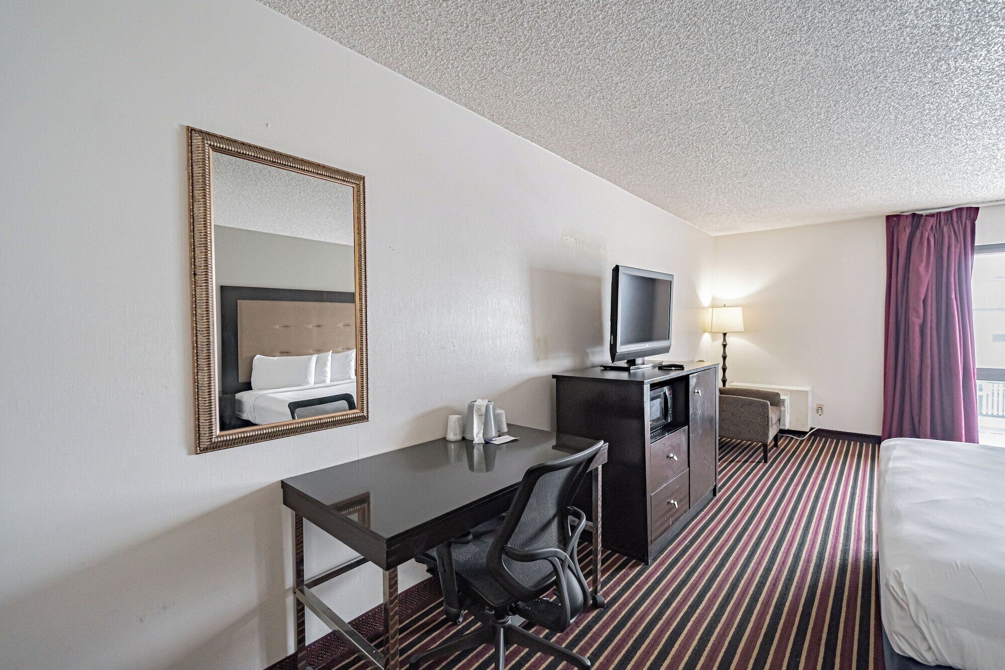 A hotel room with a bed, a desk, a chair, a television, a lamp, and a window with curtains.