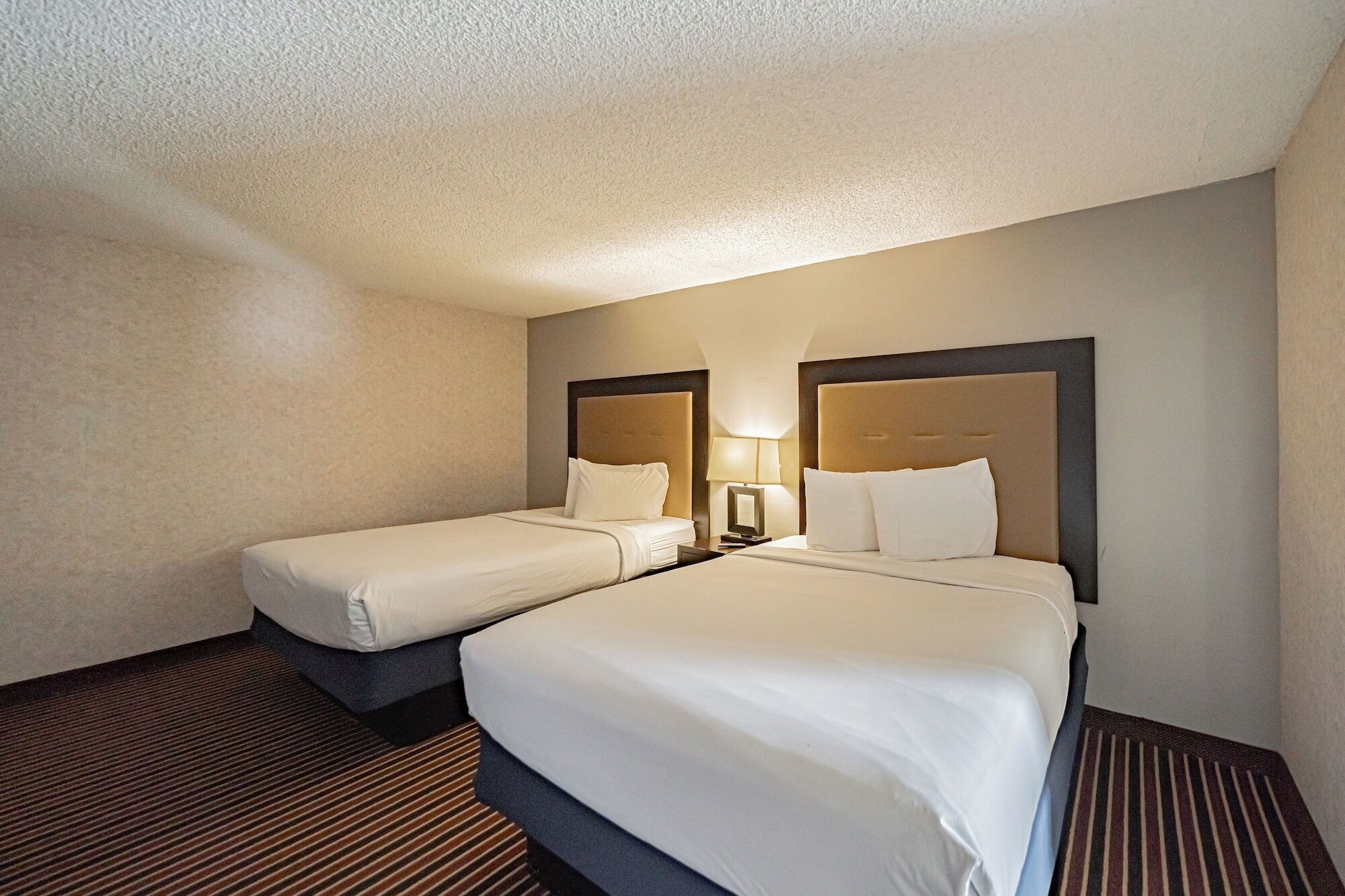 A hotel room with two beds, a striped carpet, and a lamp on the nightstand.