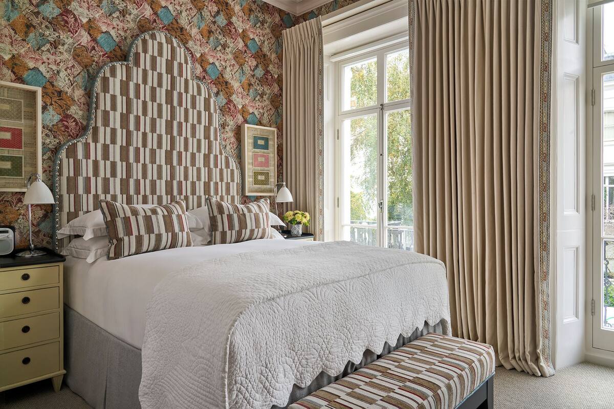 A neatly made bed with a quilted headboard, a bedside table with a lamp, and a window with curtains.