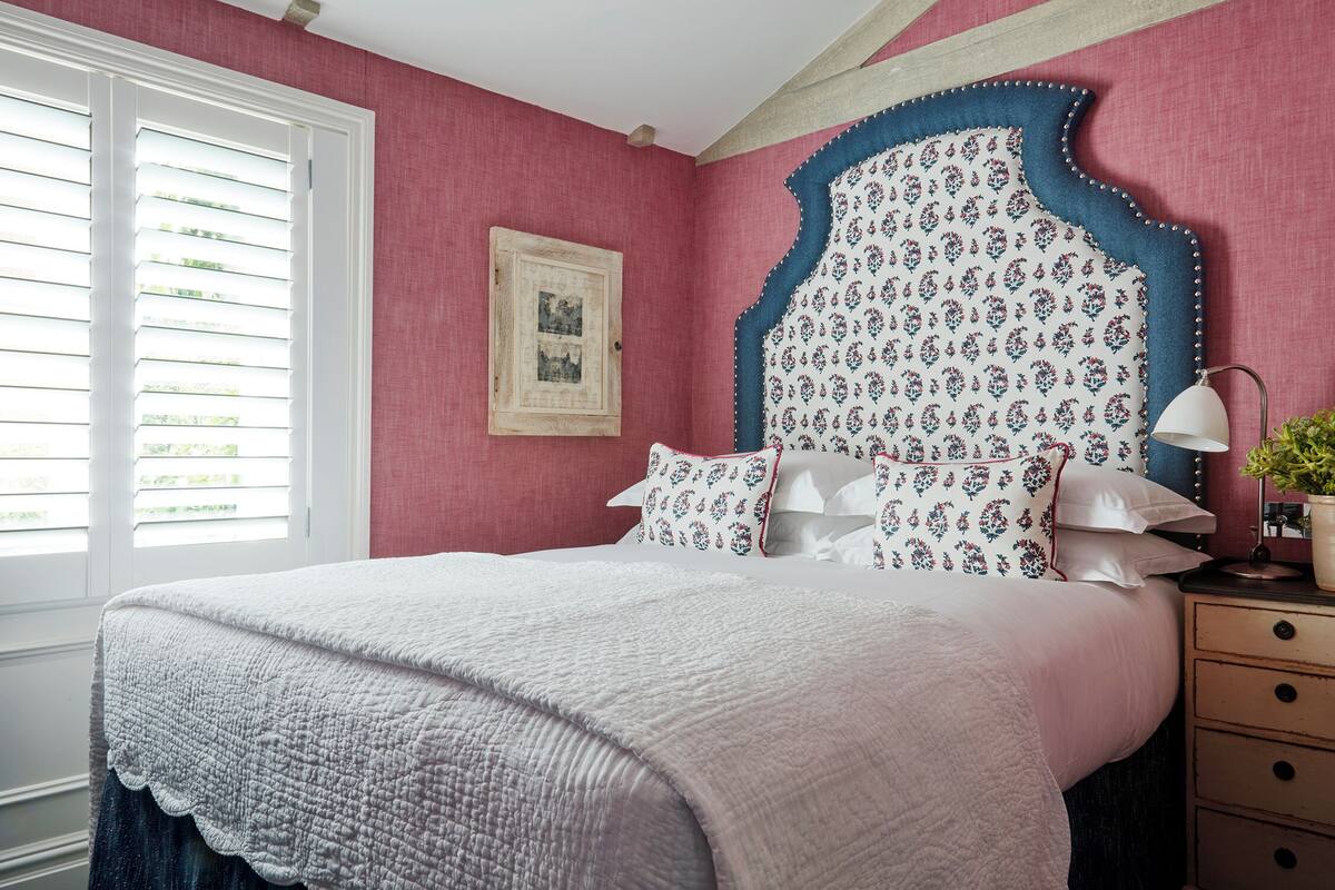 A bedroom with a large bed, a headboard with a floral pattern, a bedside table with a lamp, and a framed picture on the wall.