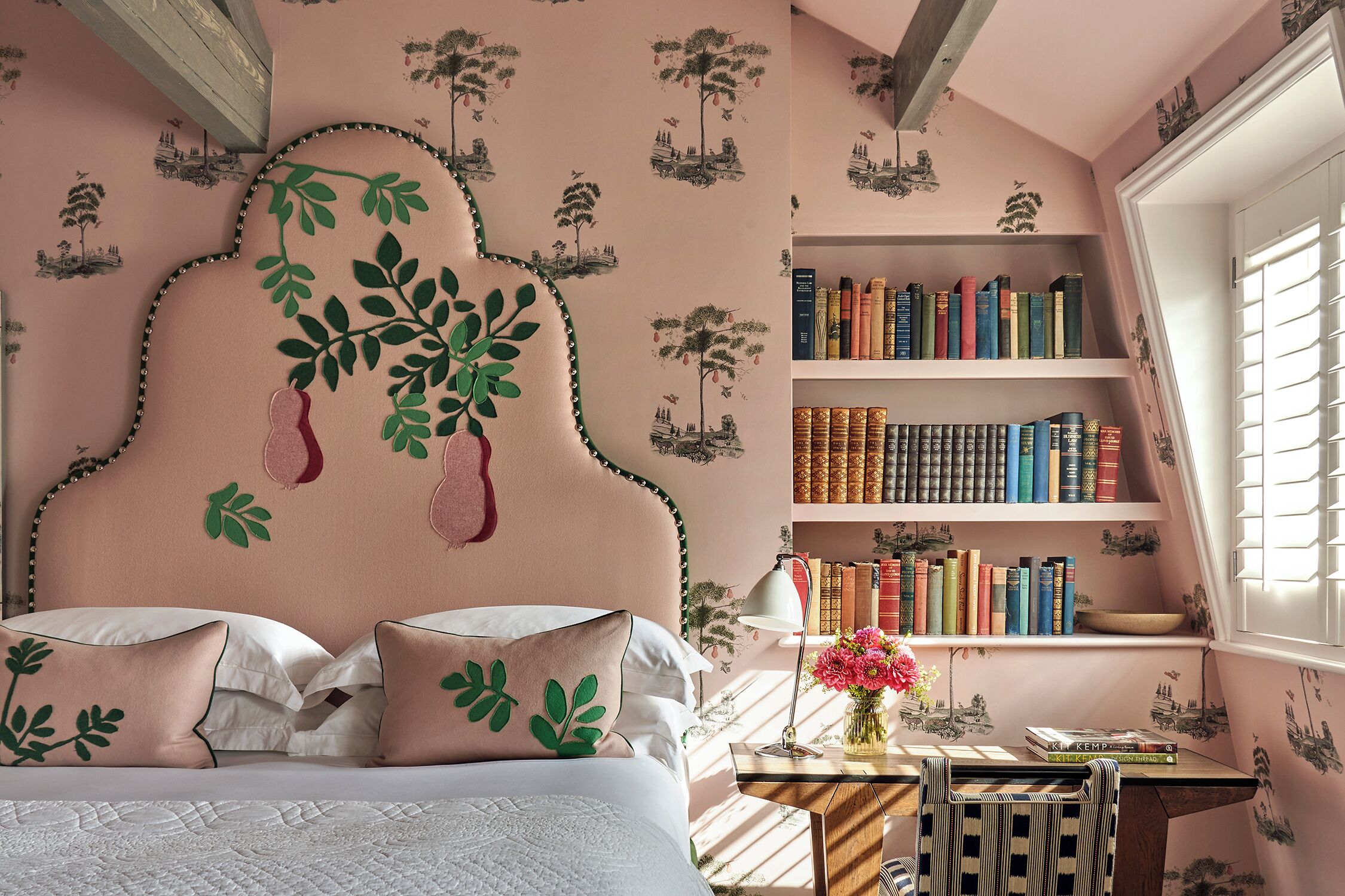 A bedroom with a bed, pillows, a bedside table with a lamp, a bookshelf, and a window with blinds.
