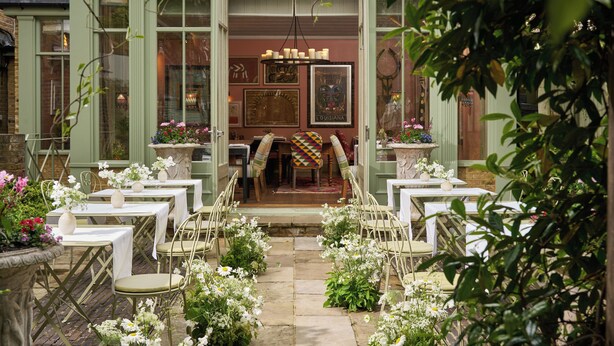Un área de comedor al aire libre con mesas y sillas dispuestas en semicírculo, rodeada de macetas con plantas y flores.