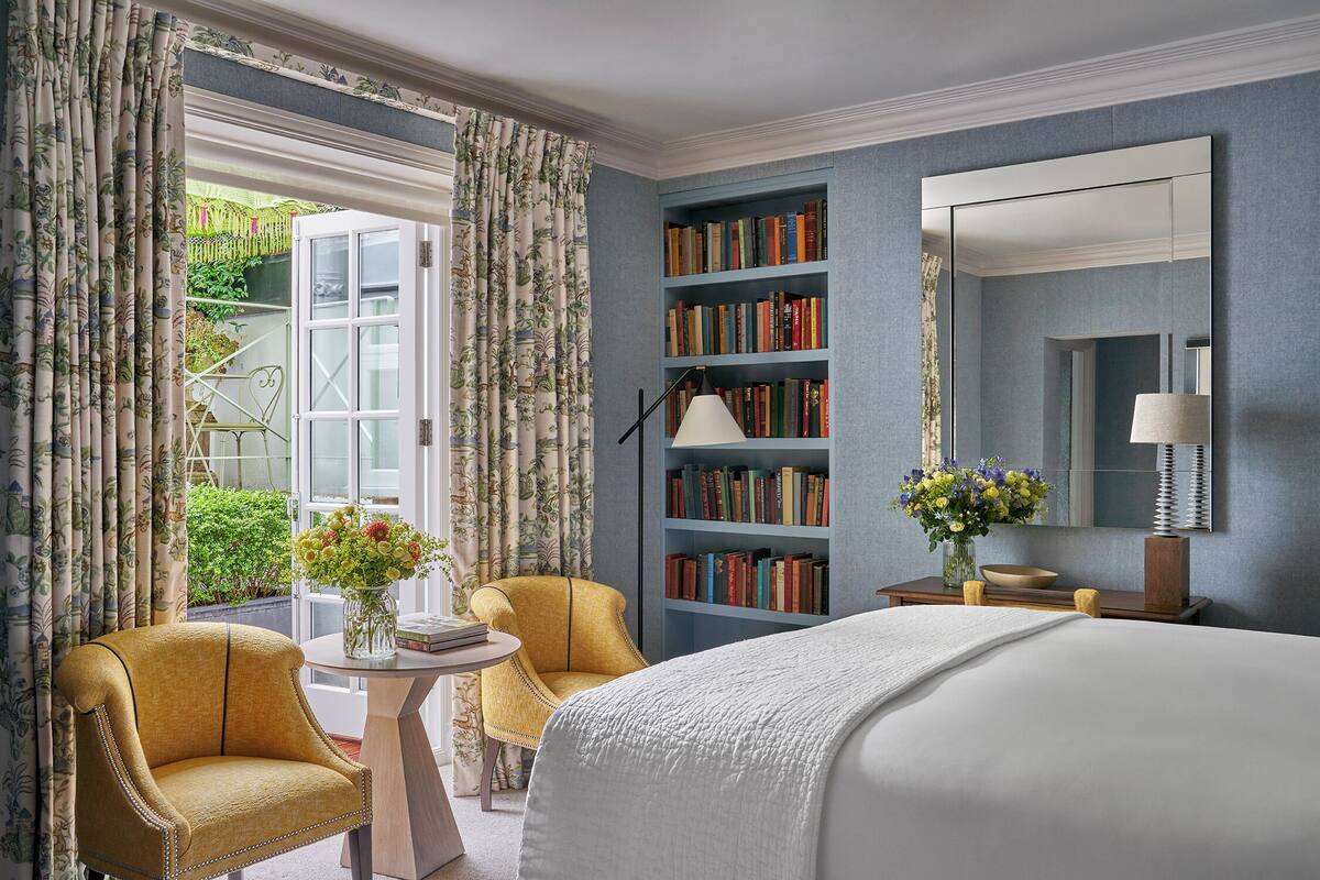 A hotel room with a bed, two armchairs, a small table with a vase of flowers, a bookshelf, and a large mirror.