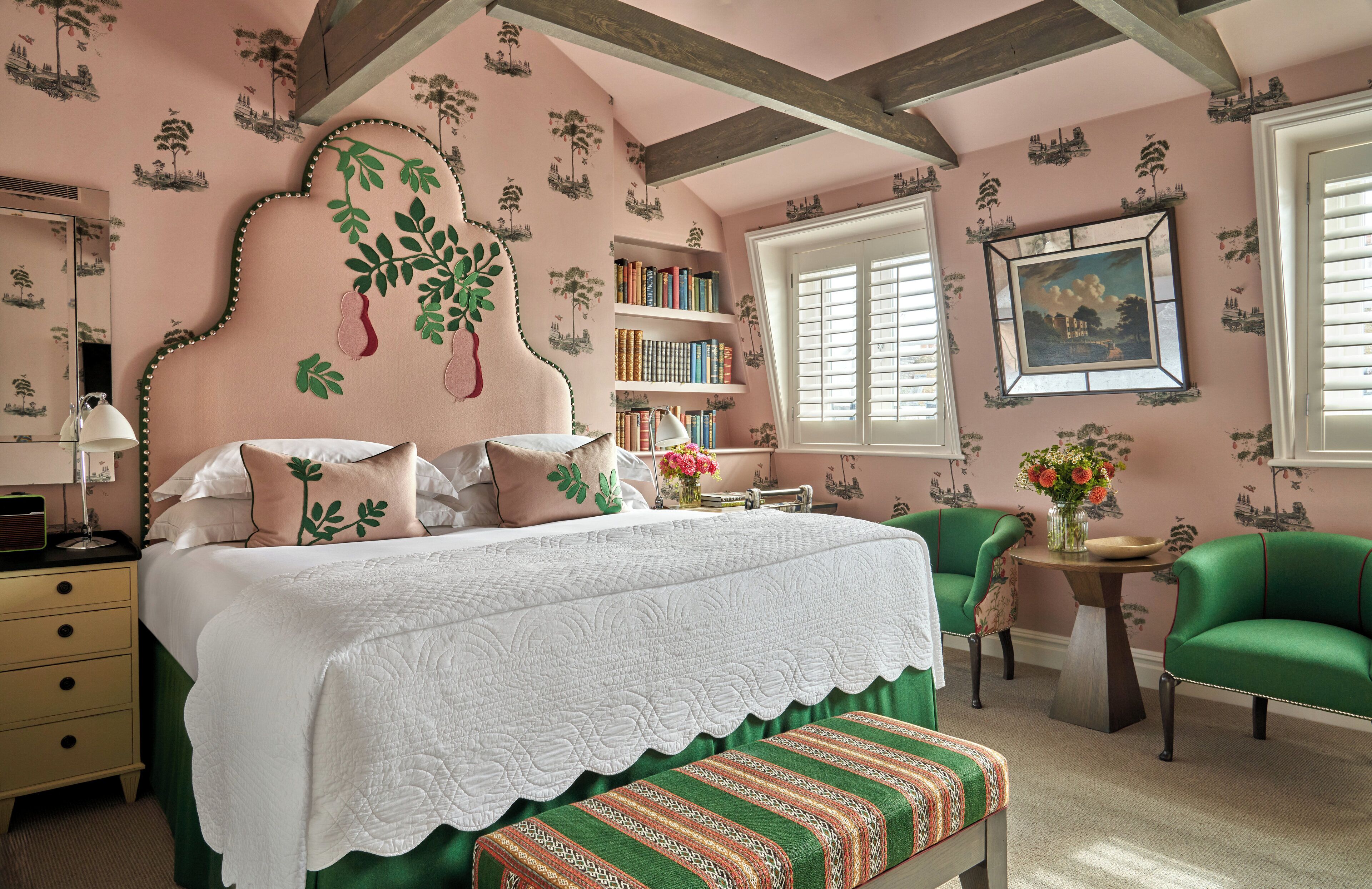 A bedroom with a large bed, a bedside table, a green armchair, a bookshelf, and a window with shutters.