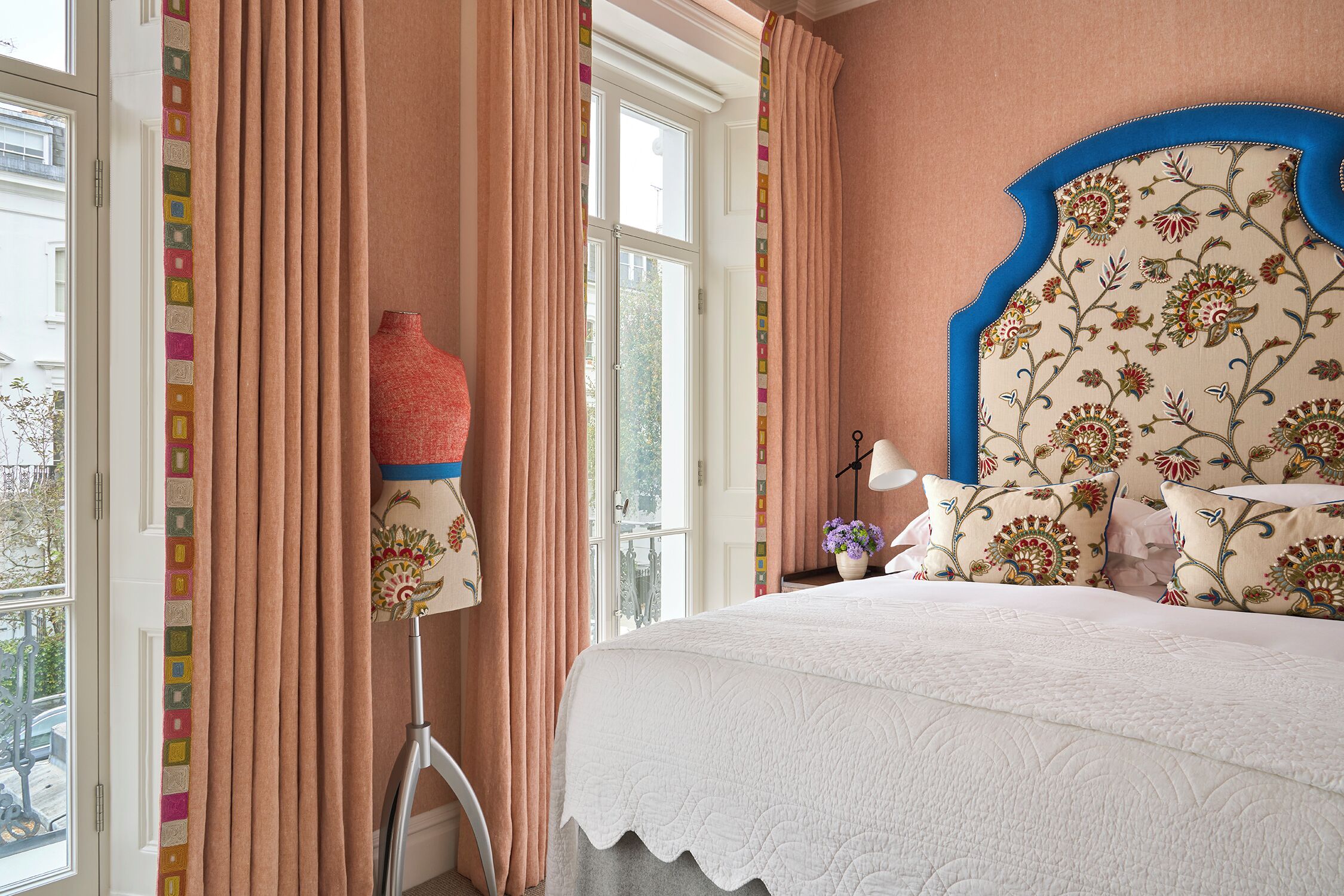 A bedroom with a large bed, a window with curtains, a bedside lamp, and a floral-patterned headboard.