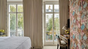 Habitación de hotel con una cama grande, un escritorio con lámpara y vistas de la ciudad a través de amplios ventanales.