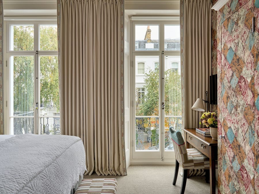 Habitación de hotel con una cama grande, un escritorio con lámpara y vistas de la ciudad a través de amplios ventanales.