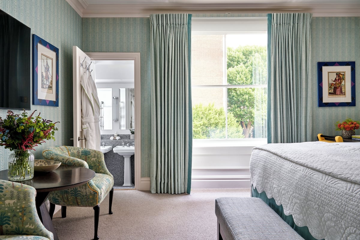 A bedroom with a bed, a window with curtains, a chair, a table with a vase, and a bathroom visible through an open door.