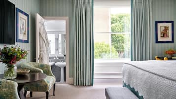 Un dormitorio con cama, ventana con cortinas, una silla, una mesa con un florero y un baño visible a través de una puerta abierta.