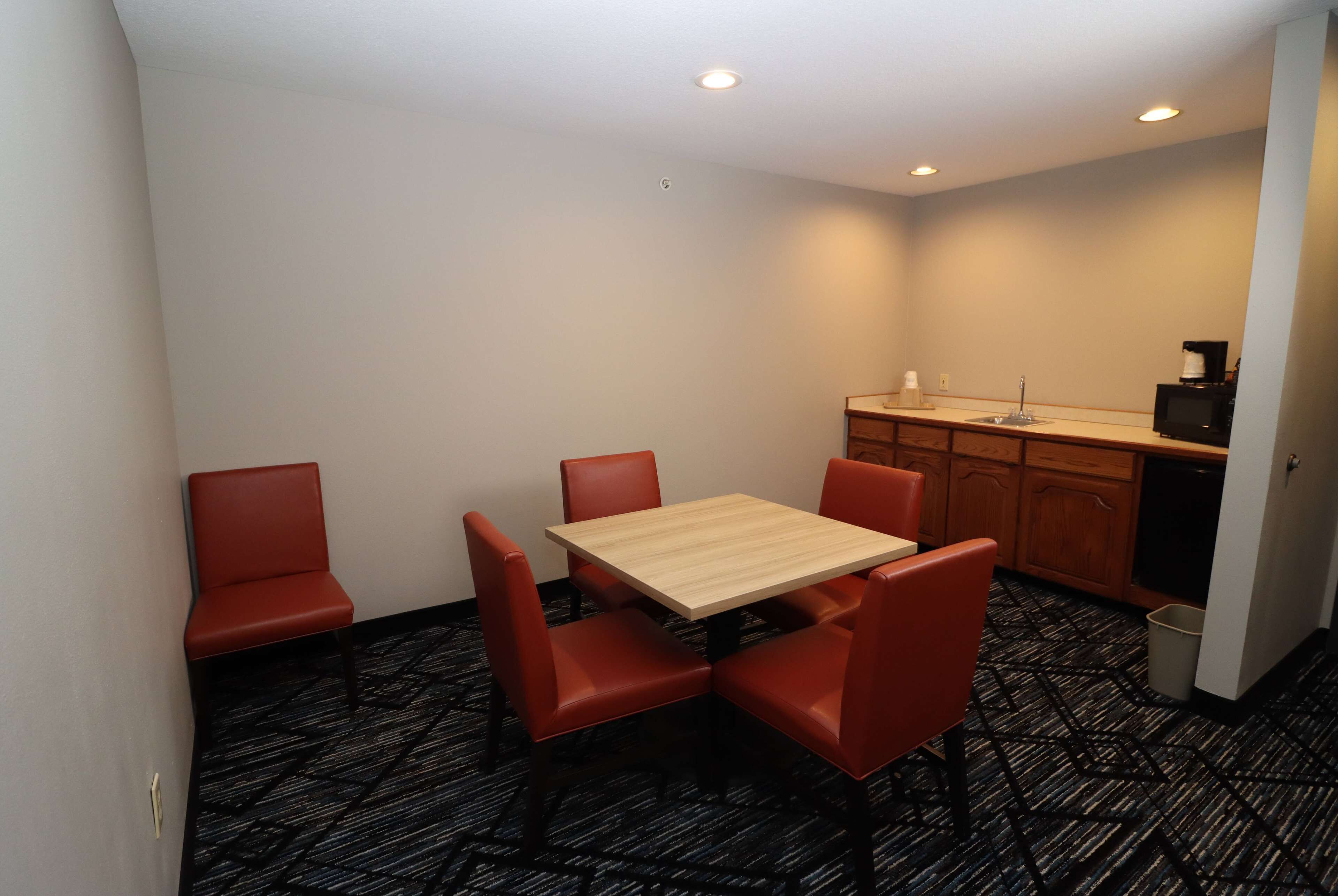 Una habitación pequeña y funcional con una mesa de madera y cuatro sillas rojas, una kitchenette con un lavamanos y una cafetera, y una alfombra estampada.