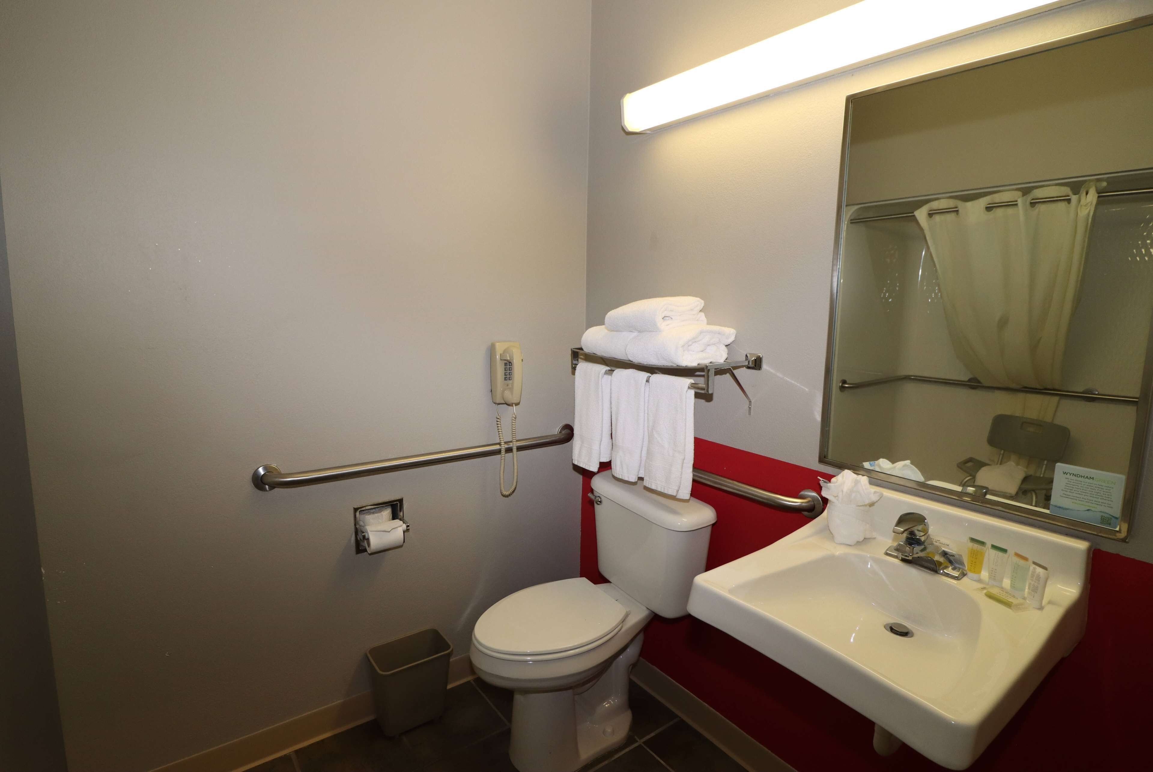 A bathroom with a toilet, sink, and a mirror. There is a handrail and a trash bin.