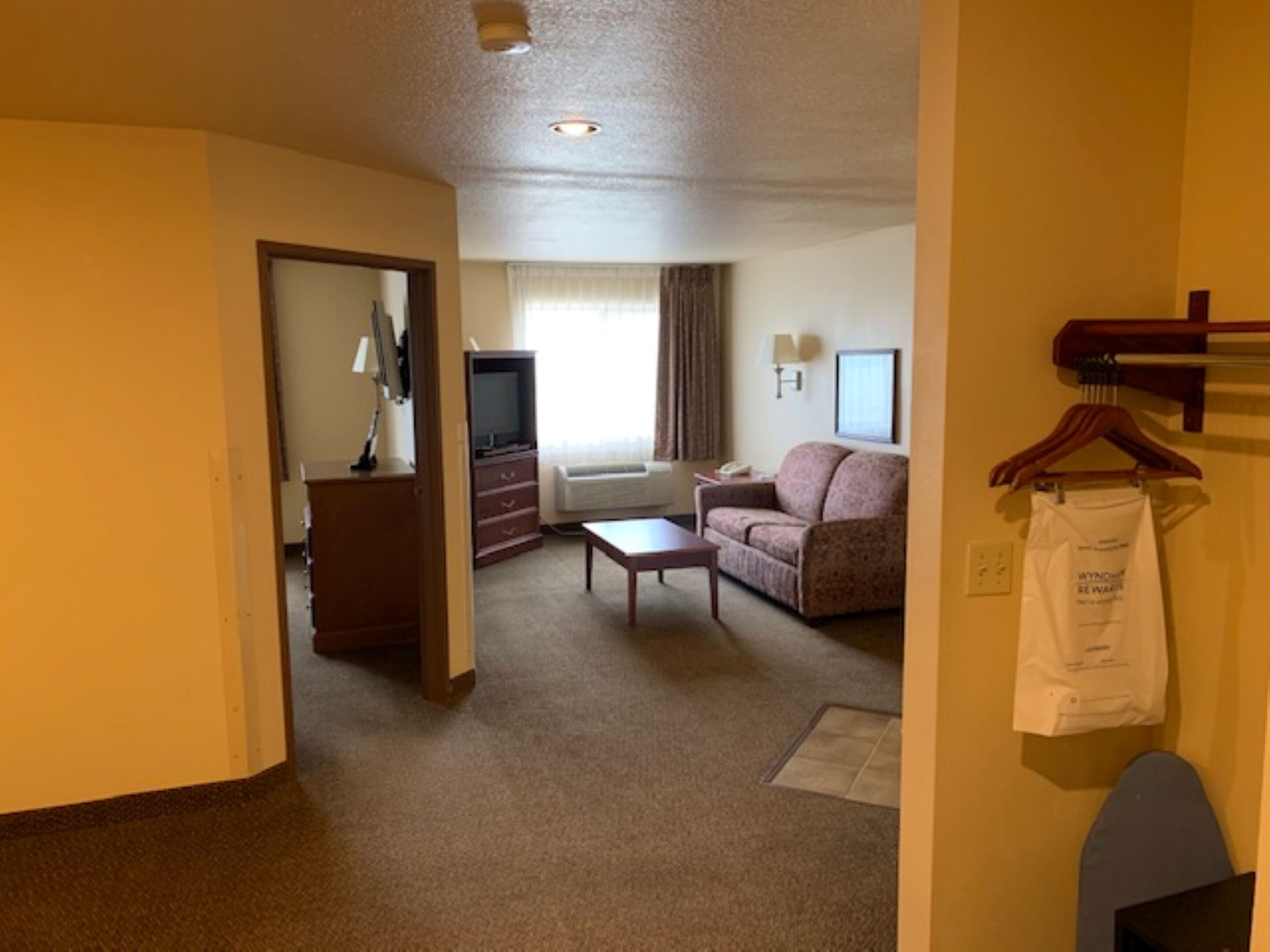 Habitación de hotel con sofá, una mesita, televisor, escritorio con lámpara y perchero.