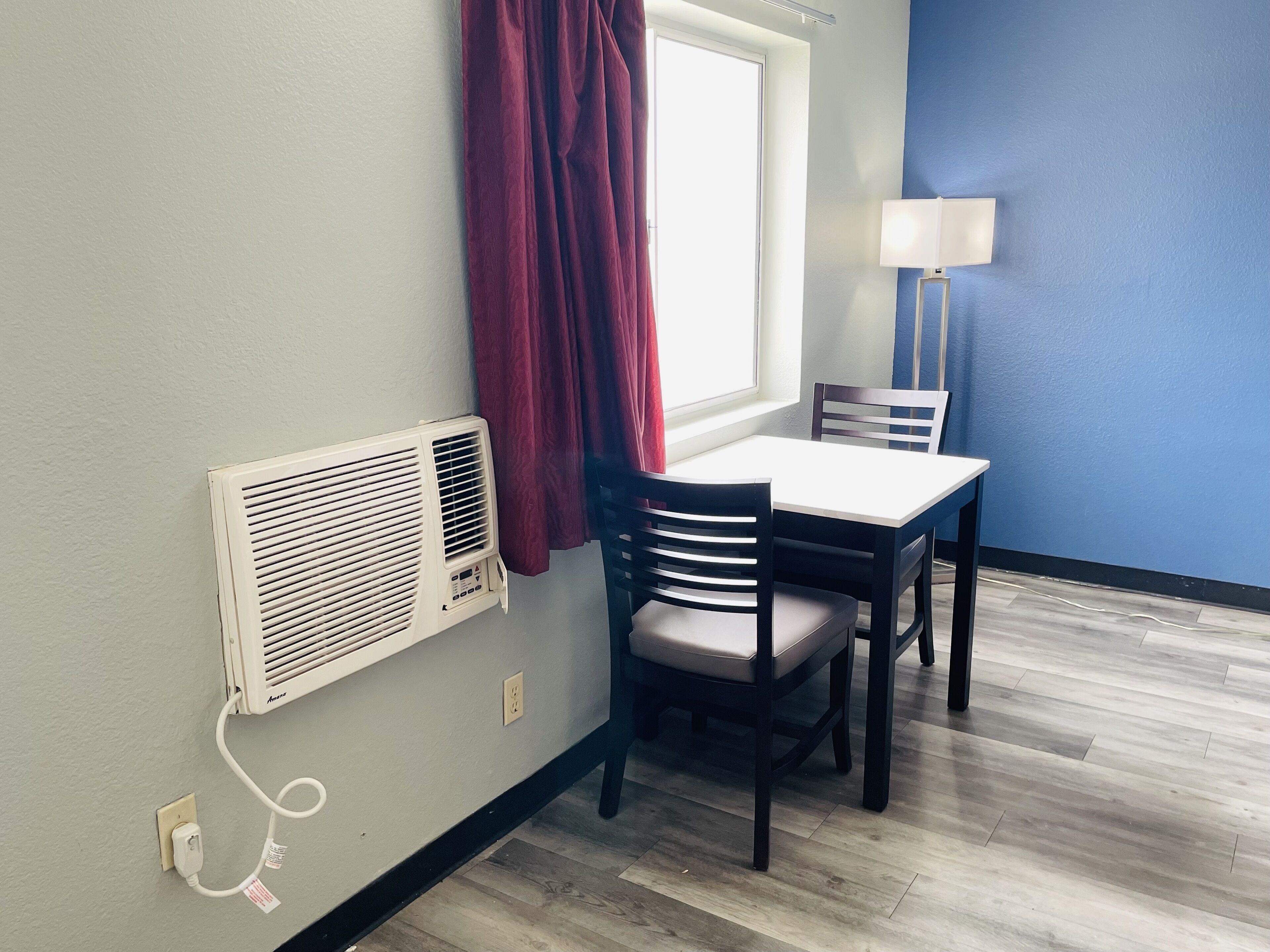 A room with a table and chairs, a window with curtains, a wall-mounted air conditioner, and a lamp.