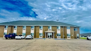 A multi-story building with a flat roof, a central entrance with a door, and a parking lot with several vehicles. - Four Seasons Inn (Bottineau)