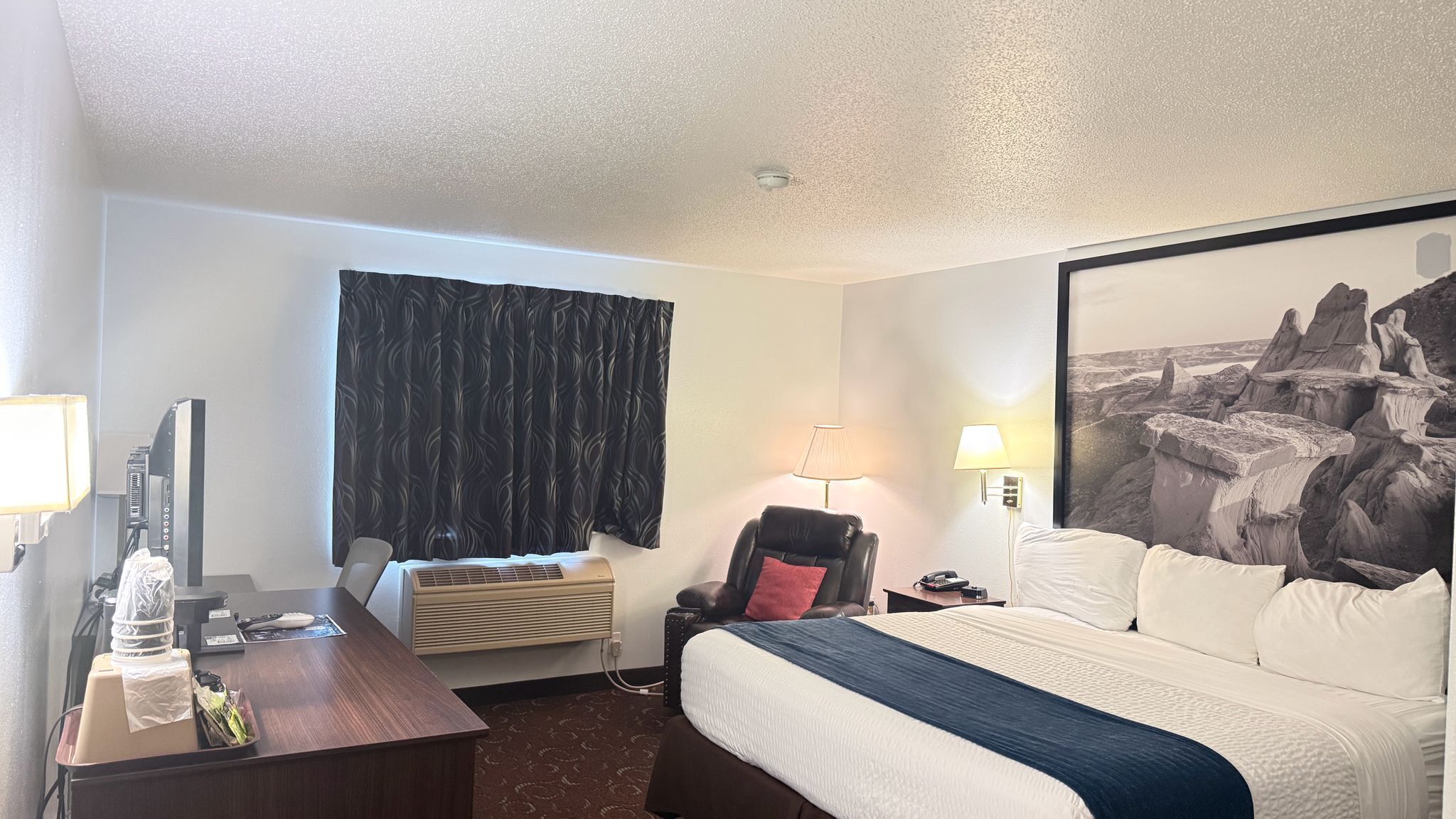 Habitación de hotel con cama, escritorio con computadora, una silla, lámpara y un cuadro grande en la pared.