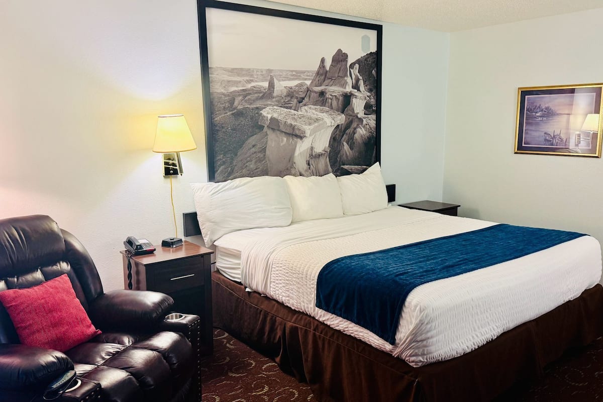 Quarto de hotel com cama, mesa de cabeceira com abajur, poltrona de couro e uma grande moldura com pintura na parede.