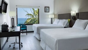 A hotel room with two beds, a desk with a chair, a TV, and a window with a view of the ocean and palm trees.