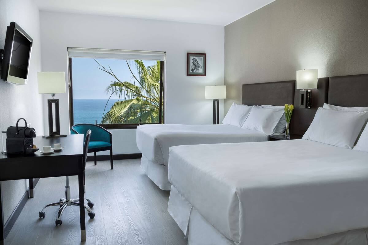 Habitación de hotel con dos camas, un escritorio con silla, televisor y ventana con vista al océano y palmeras.