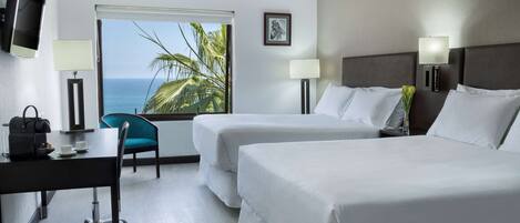 A hotel room with two beds, a desk with a chair, a TV, and a window with a view of the ocean and palm trees.