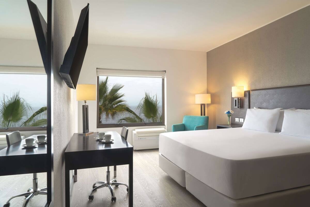 Habitación de hotel con cama, escritorio, silla, lámpara y vistas a palmeras.