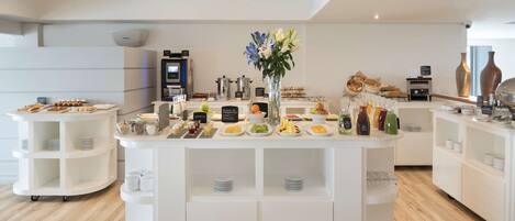 Un área de buffet moderna y bien iluminada con diversas opciones de comida, como frutas, quesos y pasteles, dispuestas en estantes y mostradores blancos.