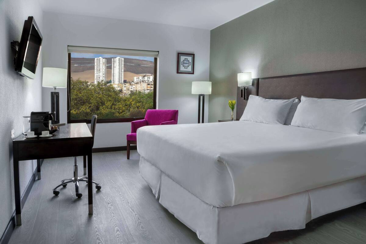 Habitación de hotel con una cama grande, un escritorio con computadora, una silla y dos lámparas. Se ve un ventanal con vista a edificios y árboles.