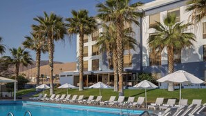 A swimming pool with lounge chairs and umbrellas, surrounded by palm trees and a building with multiple windows.
