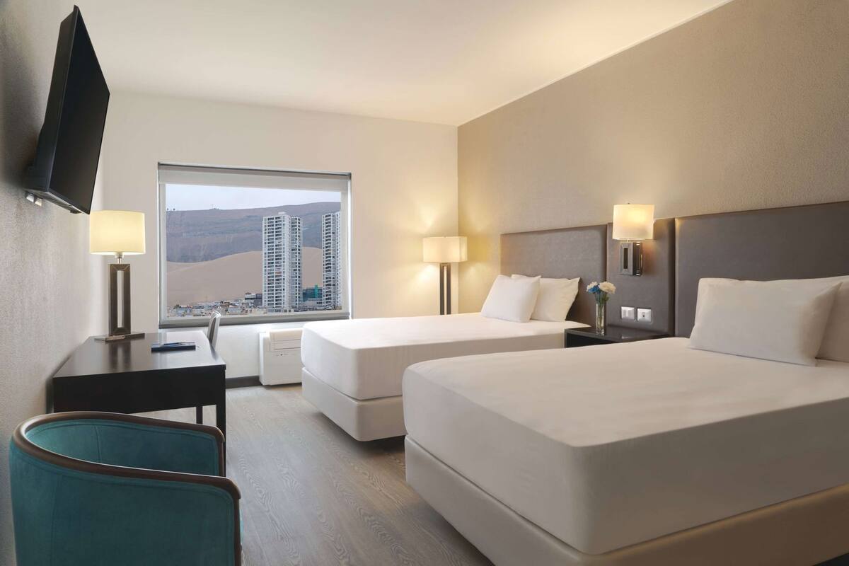 Habitación de hotel con dos camas, un escritorio, una silla, un televisor y una ventana con vista a la ciudad.