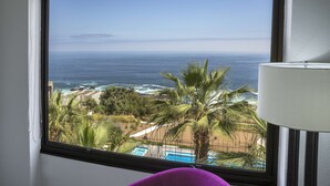 A room with a large window overlooking a beach and ocean, a pool, and palm trees.