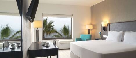 Habitación de hotel con una cama grande, un escritorio con silla, una mesita, una lámpara y vistas a palmeras a través de la ventana.