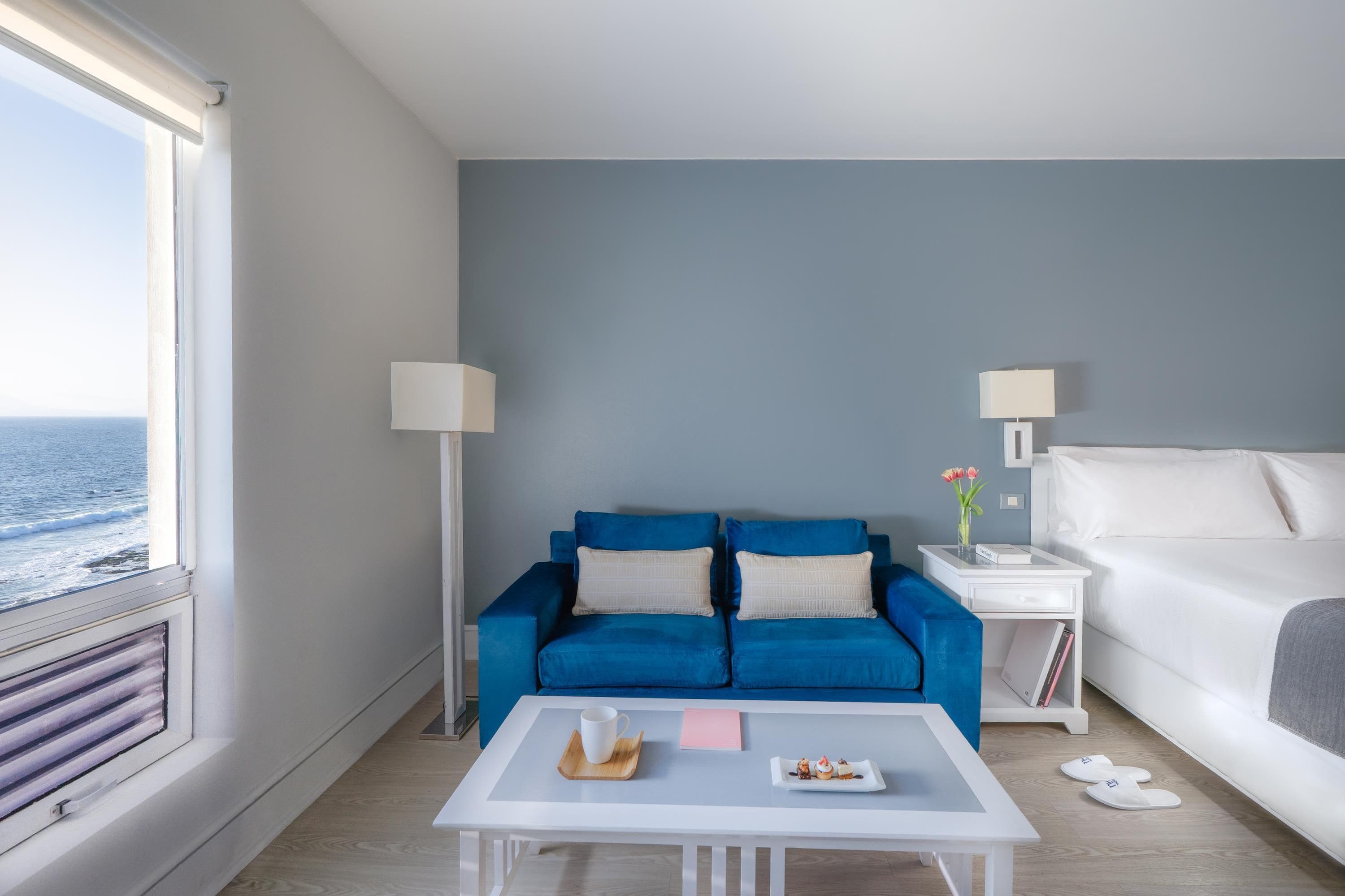Habitación de hotel con sofá azul, una mesa de centro blanca, cama y vista al mar.