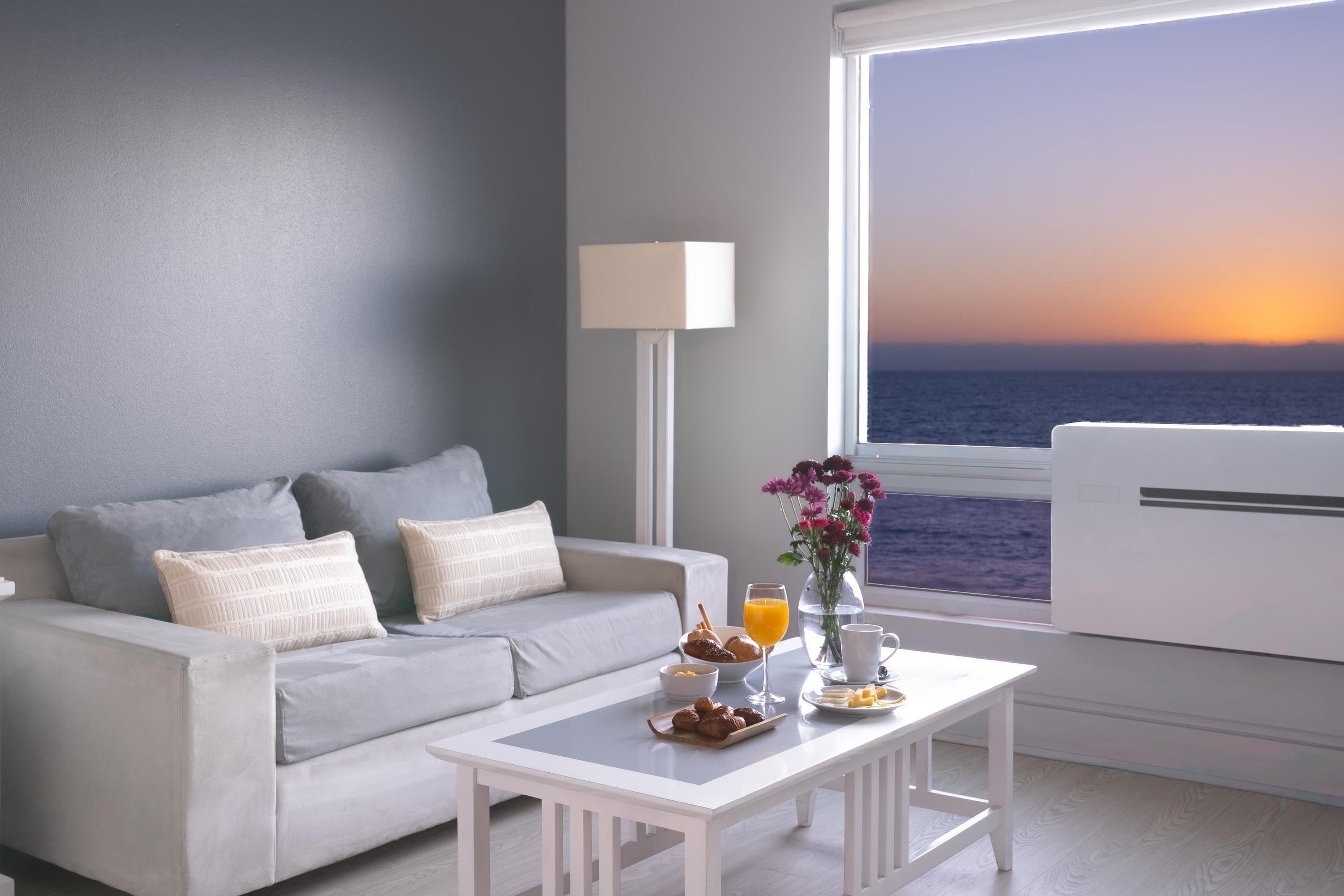 A modern living room with a grey sofa, a white coffee table with breakfast items, a lamp, and a view of the ocean at sunset.