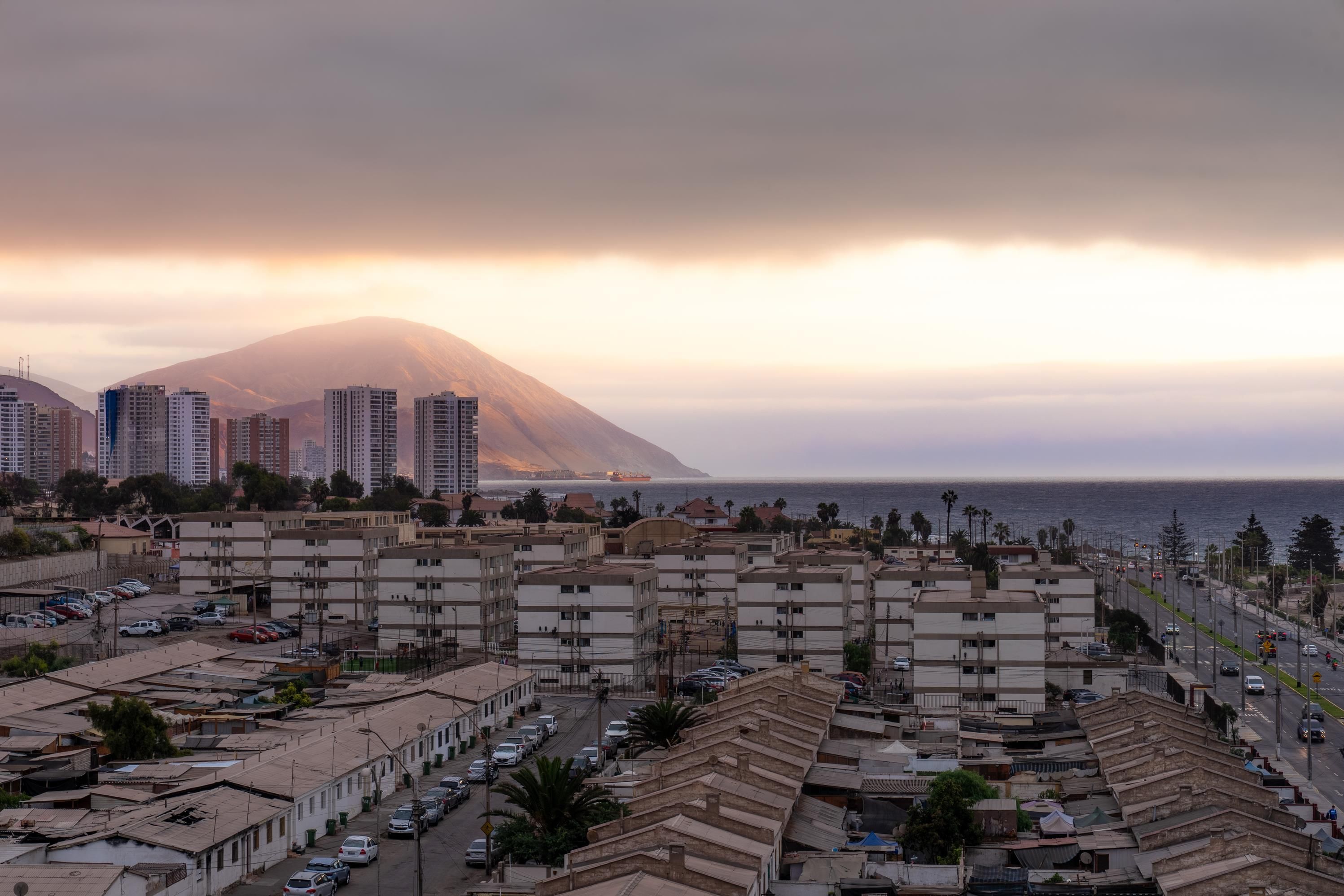 Un paisaje costero con edificios residenciales, una montaña al fondo y vista al mar.