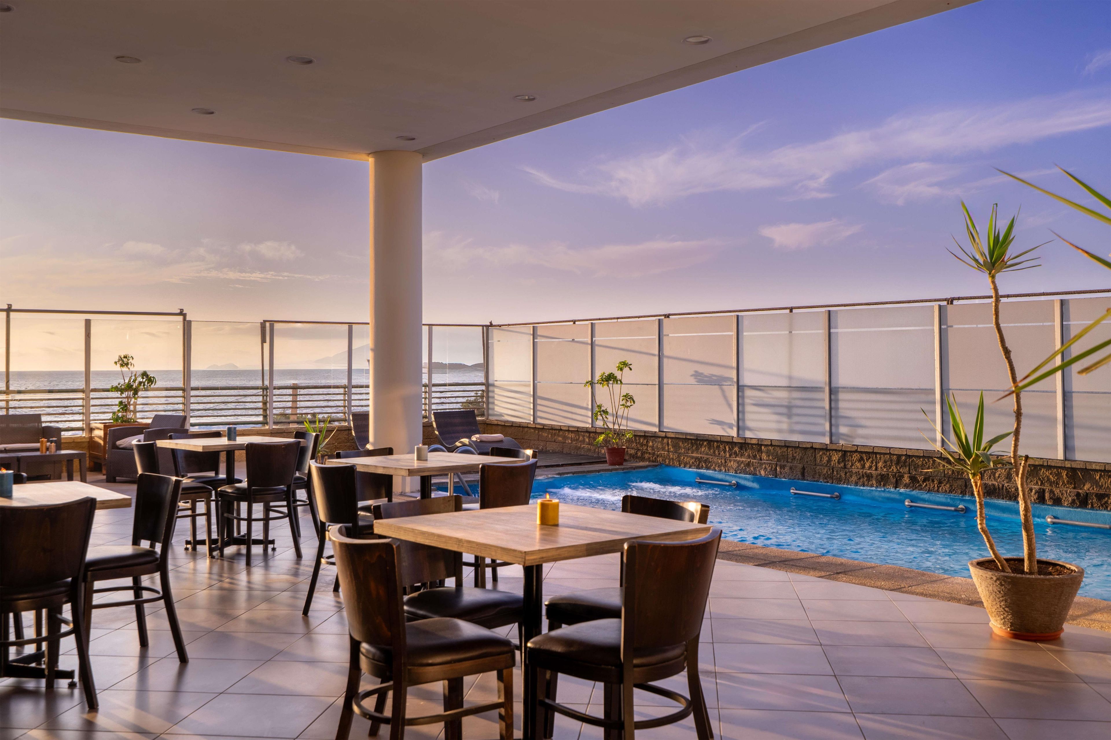 An outdoor seating area with tables and chairs, overlooking a pool and the ocean.