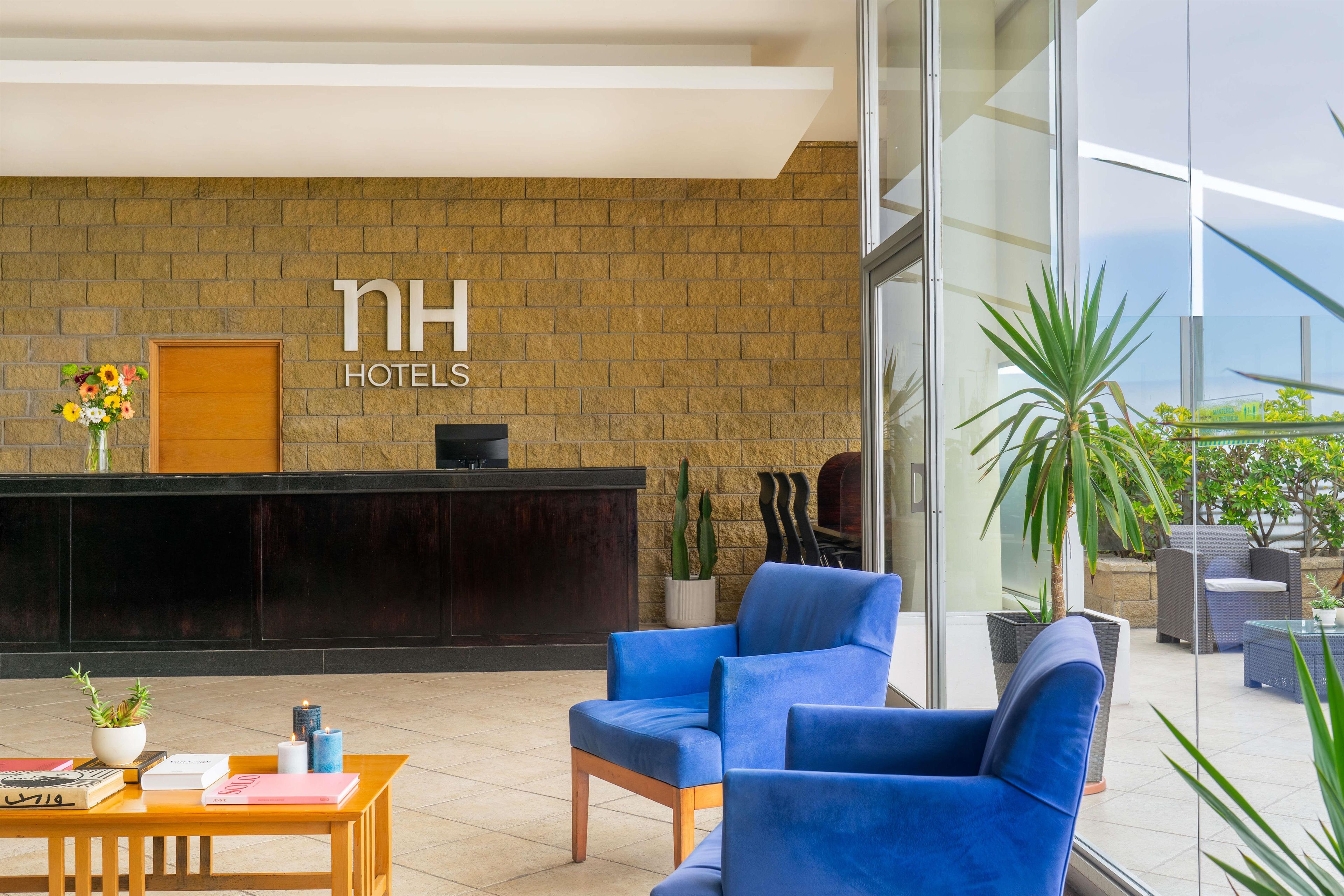 Zona de recepción de un hotel con pared de ladrillo, mostrador de recepción, sillas azules y una planta en maceta.