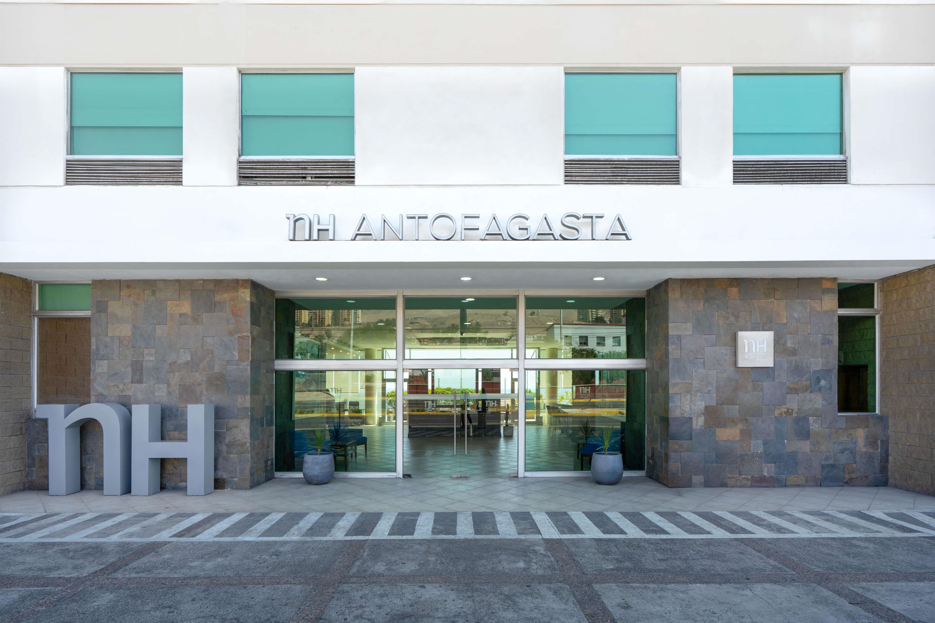 La entrada del hotel NH Antofagasta, con fachada de vidrio transparente y revestimiento de piedra, y un cartel.