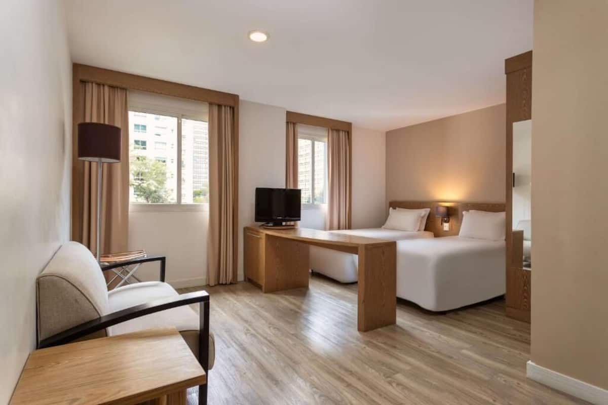 Une chambre d’hôtel avec un lit, un bureau, une chaise, une télévision et une fenêtre avec des rideaux.