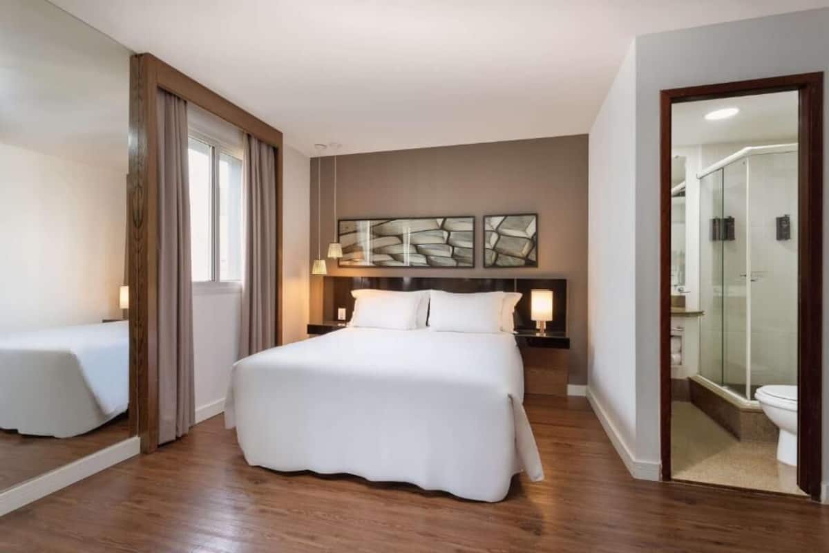 Une chambre d’hôtel avec un grand lit, une table de chevet, un miroir et une salle de bain visible à travers une porte ouverte.