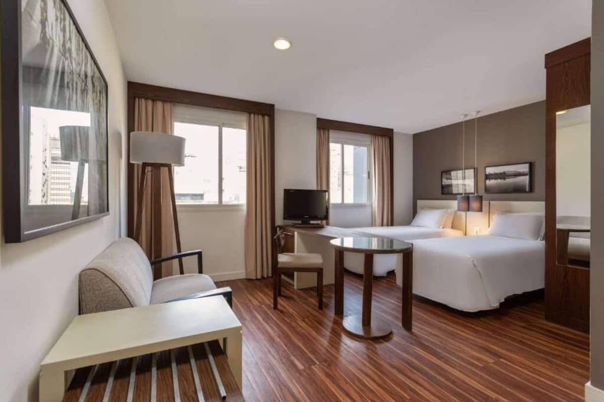 Une chambre d’hôtel avec un lit, un canapé, un bureau, une télévision et une fenêtre avec des rideaux.