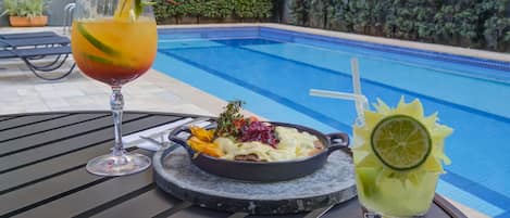A dining table with a meal and two drinks by a poolside.
