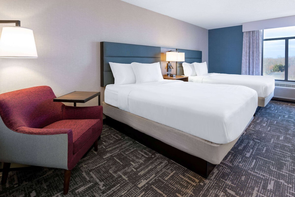 A hotel room with two beds, a red armchair, a small table, and a lamp.