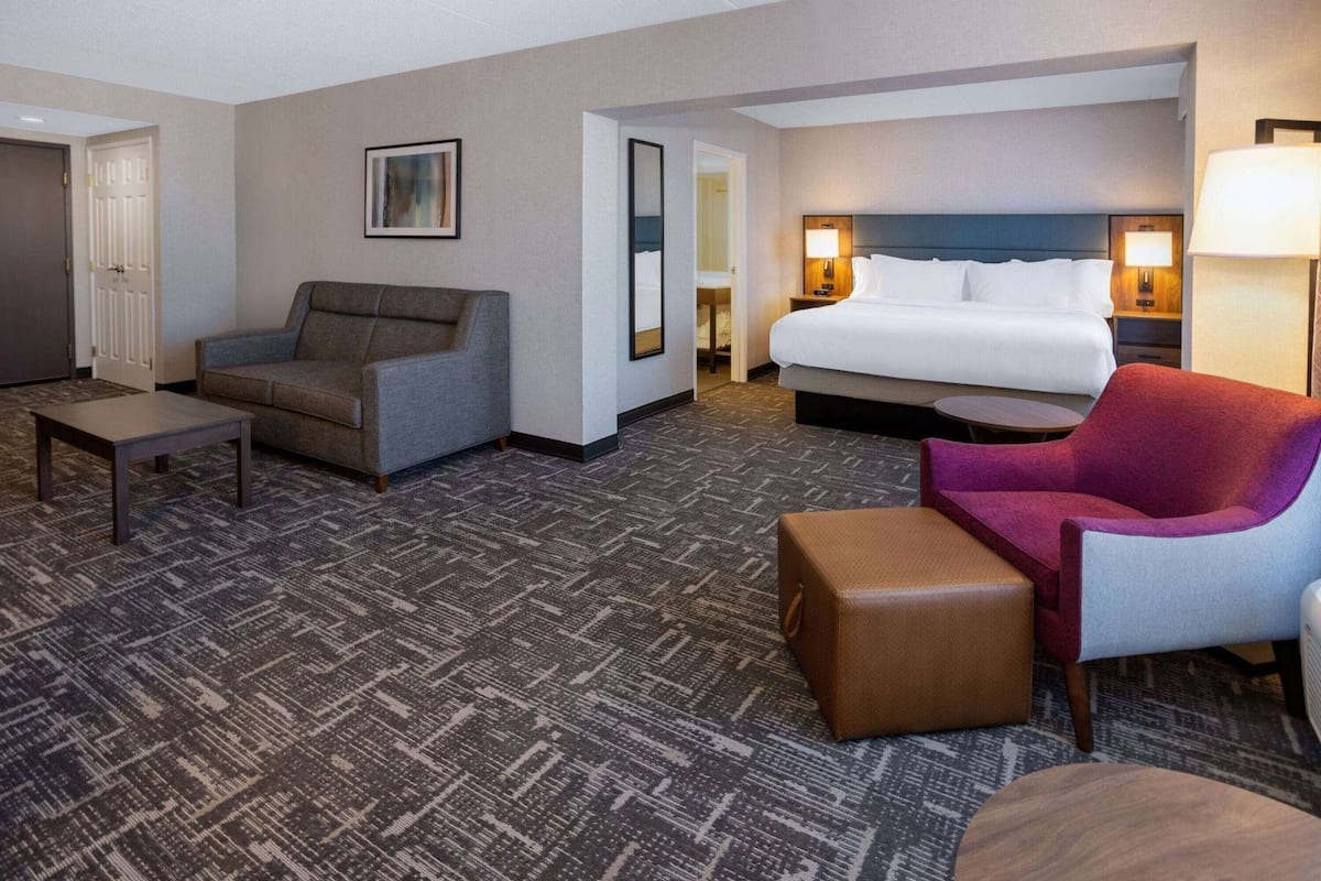 A hotel room with a bed, a grey sofa, a red armchair, a brown ottoman, a small table, and a wall-mounted TV.