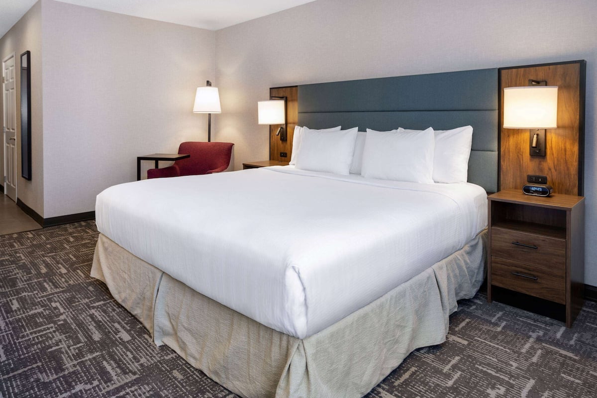 A hotel room with a large bed, two bedside tables with lamps, a red chair, and a carpeted floor.