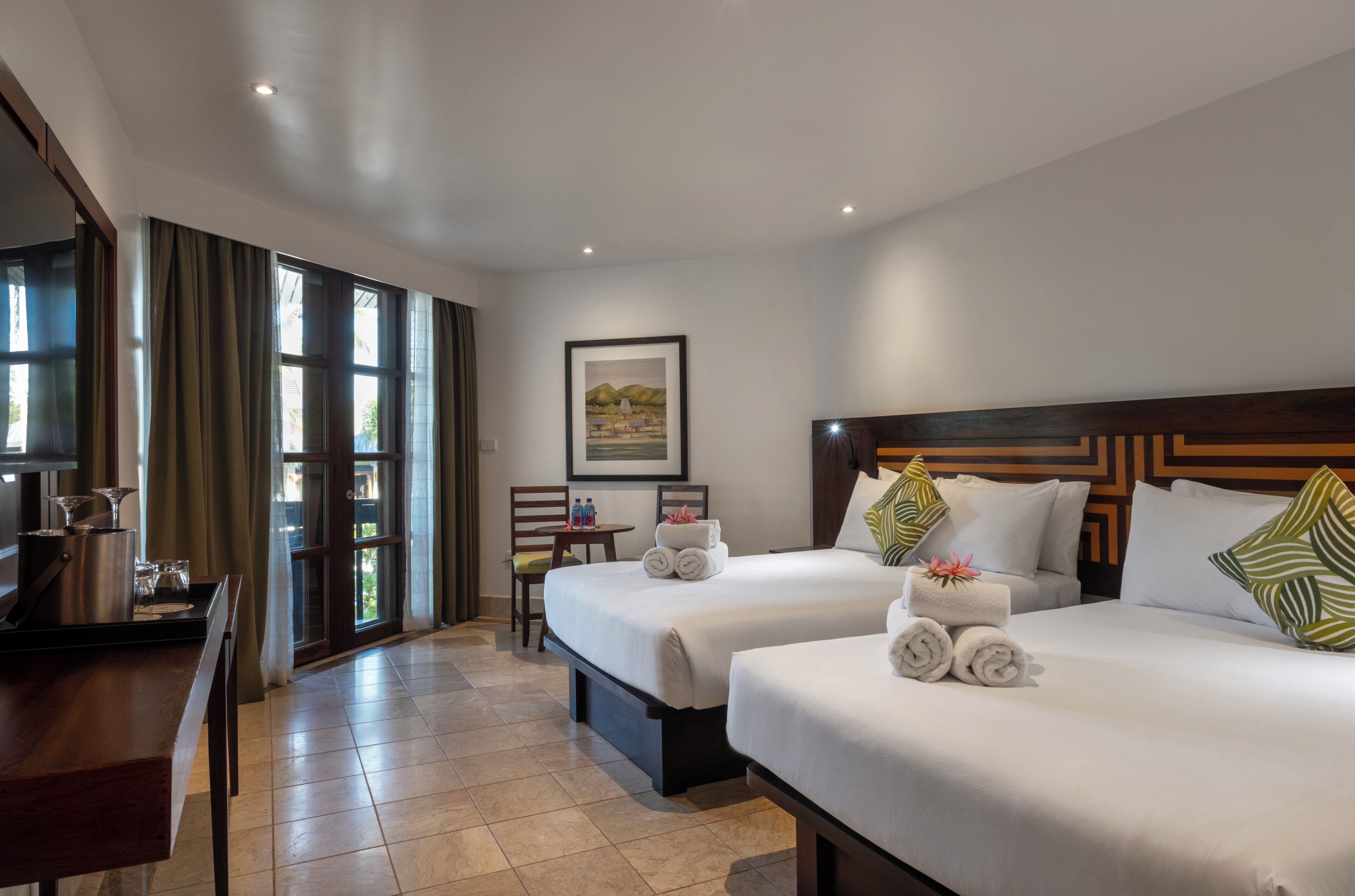 A hotel room with two beds, a desk, a chair, a table with a bottle, and a framed picture on the wall.