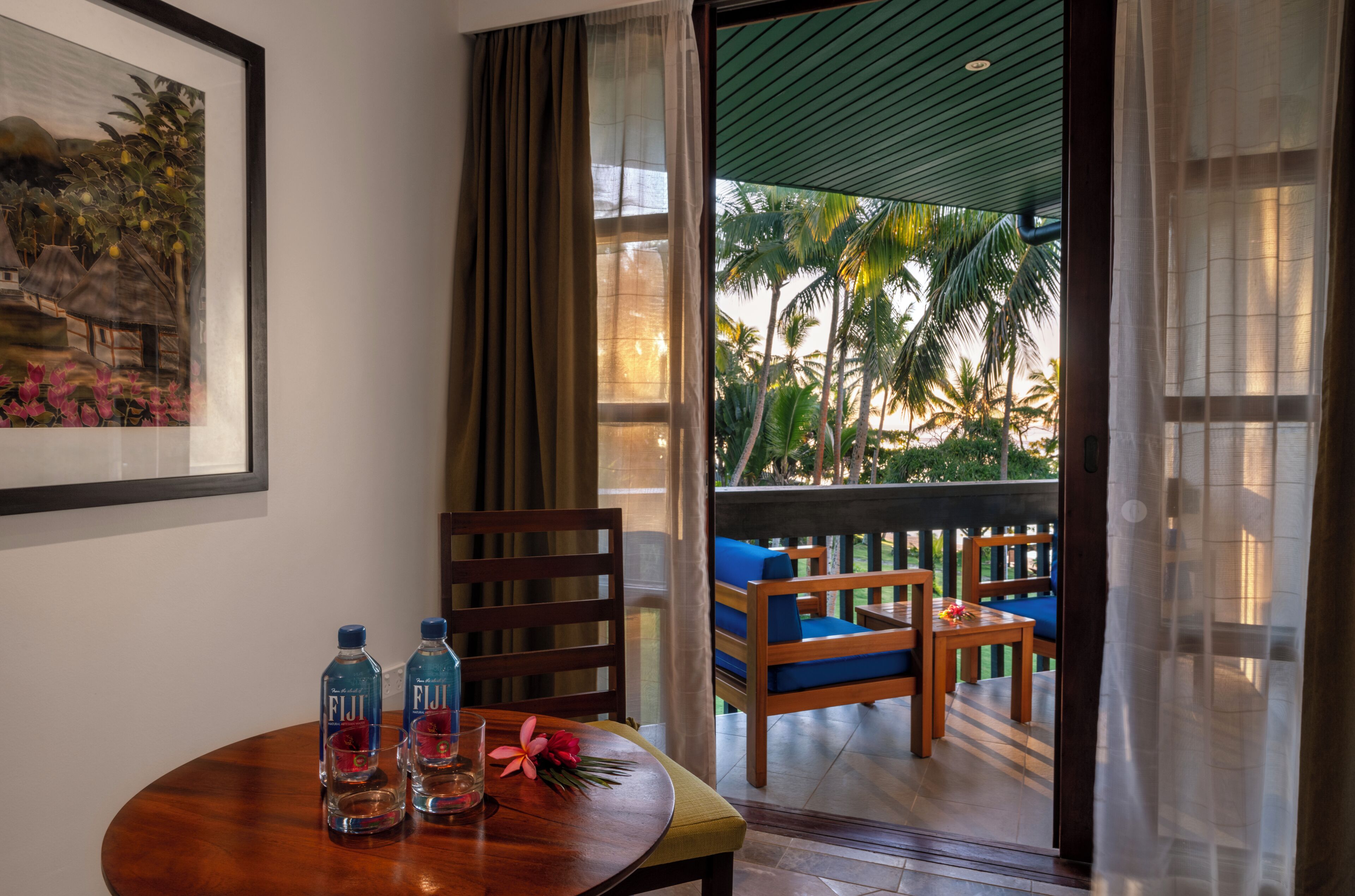 A room with a wooden table, two chairs, a sofa, and a balcony with a view of palm trees.